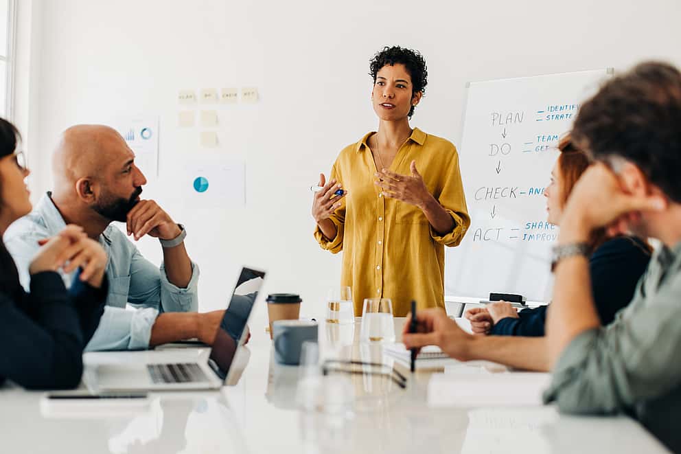 A businesswoman sitting at her desk using her phone, in a busy office environment, symbolizing grenke's commitment to helping ambitious partners grow by offering leasing solutions for sales without boundaries.